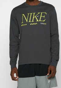 Langärmliges, dunkelgraues Nike-Shirt mit leuchtend gelber Markenkennzeichnung und Text. Hergestellt aus glattem Stoff mit Rundhalsausschnitt.