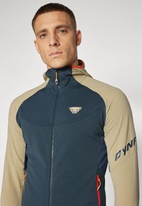 Men's zip-up jacket, featuring navy and beige fabric, with a textured finish, hood, and logo detail on the chest and sleeve.