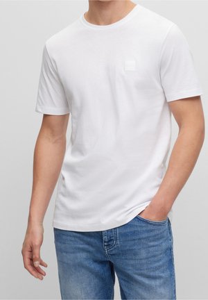 T-shirt blanc en coton à manches courtes et col rond. Présente un logo carré texturé sur le côté gauche de la poitrine. Porté avec un jean bleu.