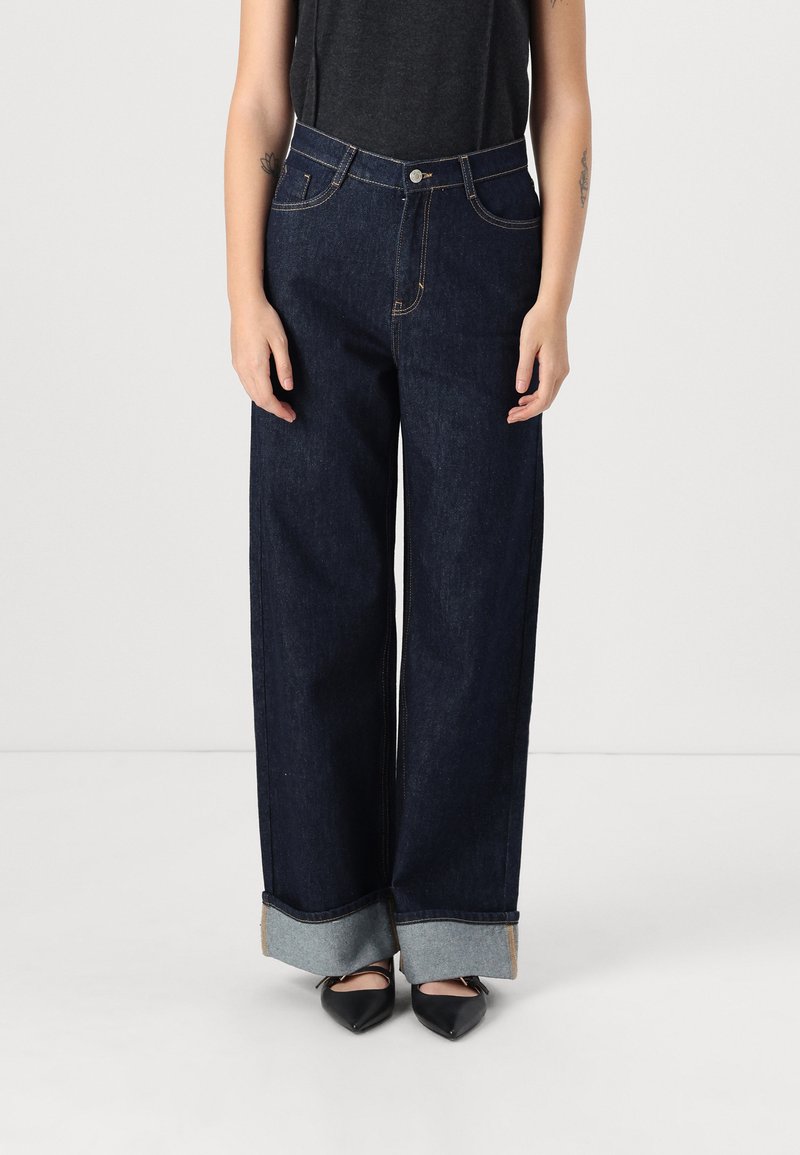 Jeans de mezclilla oscura de pierna ancha con cintura alta, que presentan puños en un azul claro contrastante y costuras doradas, combinados con zapatos negros de punta.