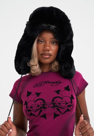 Chapeau en fausse fourrure noire avec des rabats pour les oreilles, présentant un logo brodé à l'avant. Le modèle porte une chemise violet avec un design graphique noir audacieux.