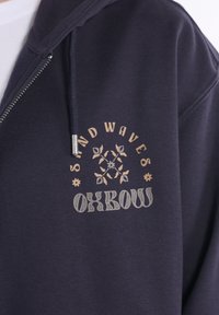 Sweat à capuche marine avec fermeture éclair, cordon de serrage et design imprimé en beige et gris avec le texte "SAND WAVES OXBOW" et des motifs floraux.