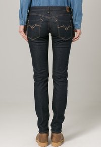 Jeans de denim oscuro con un diseño ajustado, que presentan bolsillos traseros con líneas de costura y un parche de cuero en la cinturilla.
