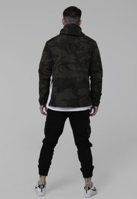 Mand med nakketatovering iført sort camouflaged jakke, sorte cargo bukser og hvide sneakers, stående med ryggen til imod en ensfarvet baggrund.