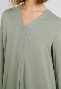 Hellgrüne Bluse aus glattem Stoff. Verfügt über einen V-Ausschnitt und lange Ärmel mit einem lockeren Schnitt. Minimale Nähdetails.