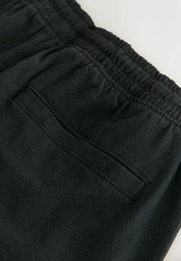 Gros plan d'un pantalon noir à taille élastique avec une poche passepoilée unique à l'arrière et des coutures visibles sur un tissu doux.