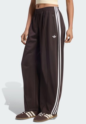 Osoba nosząca czarne szerokie spodnie dresowe Adidas z białymi paskami oraz pasujące brązowo-białe buty sportowe Adidas.