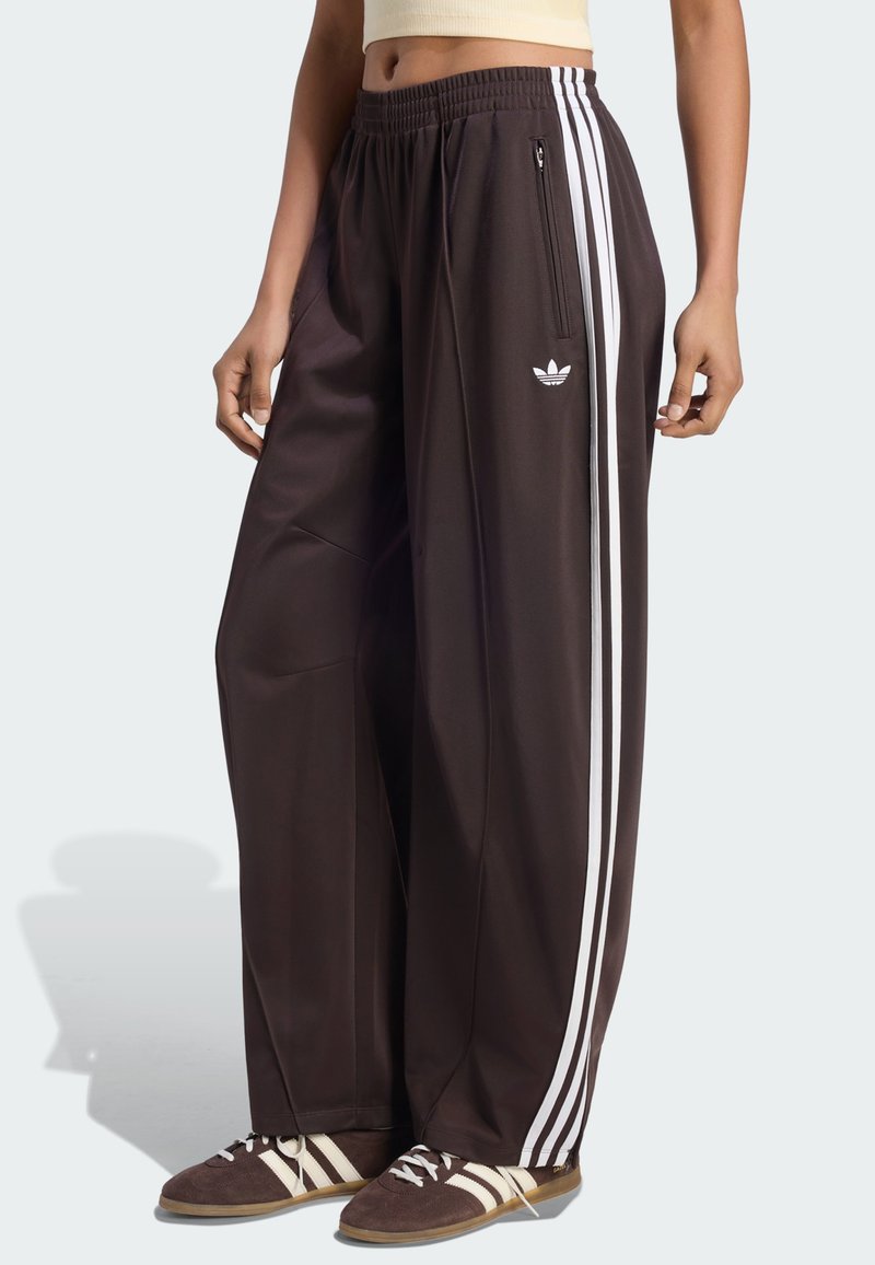 Άτομο που φοράει μαύρη φαρδιά φόρμα Adidas με άσπρες ρίγες και αντίστοιχα καφέ και άσπρα αθλητικά παπούτσια Adidas.