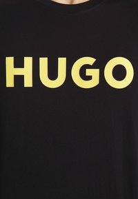 Tela negra con grandes letras amarillas en negrita que dicen "HUGO" centradas en el área del pecho.
