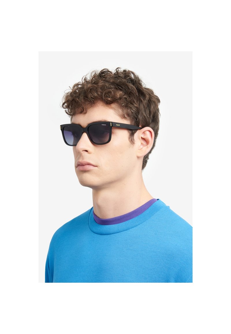 Ochelari de soare negri cu montură rectangulară, lentile cu degradé și accente aurii, purtați de un model într-un pulover albastru cu guler purpuriu.