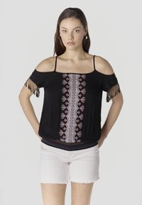 Joven de pie con las manos en los bolsillos, vestida con una blusa negra de hombros descubiertos con bordados coloridos y shorts de mezclilla blancos.