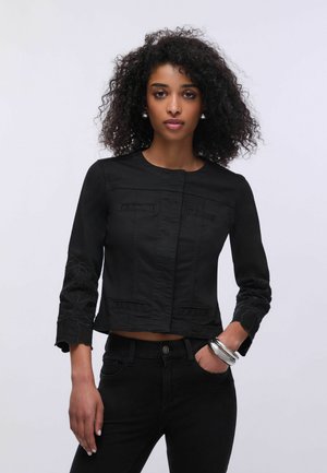 Donna con capelli ricci indossa una giacca nera aderente con ricami floreali sulle maniche e pantaloni neri, posando con una mano in tasca.