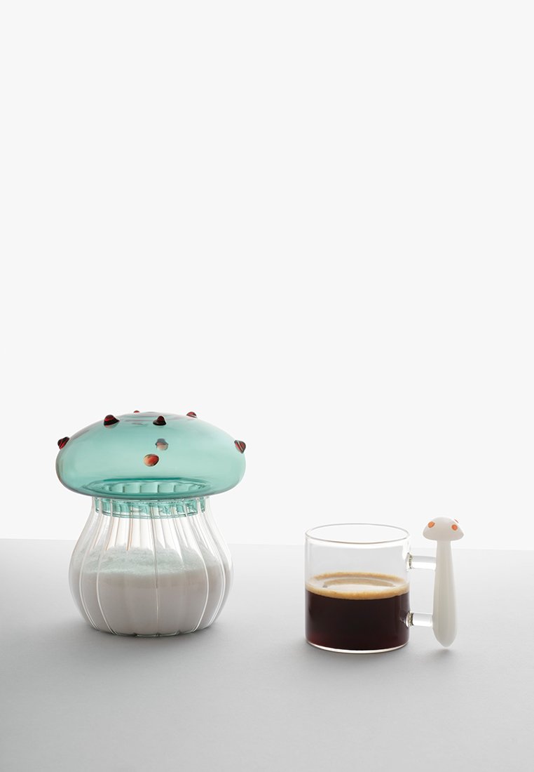 Szklany dozownik cukru z niebieską pokrywką i czerwonymi akcentami obok przezroczystej szklanki espresso, z białą rączką w kształcie grzybka.