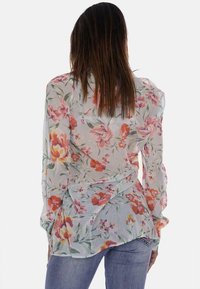 Blusa floreale in tessuto trasparente con base azzurro chiaro, caratterizzata da fiori rosa e arancioni, maniche lunghe e un design a volant sul retro.