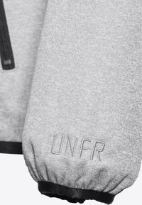 Lys grå stoffett med elastisk svart mansjett og "UNFR" brodert nær håndleddet, en del av et glidelåsgarment.
