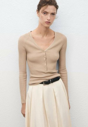Vrouw in een beige geribbelde V-hals gebuttonde trui, gestoken in een hooggetailleerde crèmekleurige plissérok met een zwarte riem.