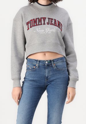 Nainen, joka pukeutunut harmaaseen lyhennettyyn collegepaitaan, jossa lukee ”TOMMY JEANS New York”, ja korkeavyötäröisiin sinisiin farkkuihin, seisoo valkoisen taustan edessä.
