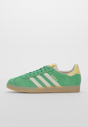 Basket Adidas Gazelle en daim vert avec des bandes blanches, un languette talonnière jaune et une semelle en gomme, vue de côté sur fond blanc.