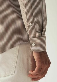 Braun-weiß gestreiftes Hemd mit einem Knopf am Ärmelbündchen. Baumwollmaterial. Nahaufnahme des Ärmels und der Hand vor hellem Hintergrund.