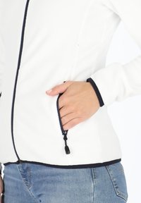 Giacca in pile bianca con zip e rifiniture blu navy. Presenta una tasca laterale e una texture morbida. Il design è aderente con una forma casual.