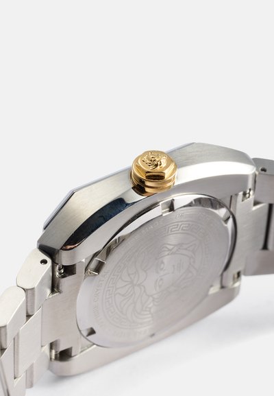 Reloj de acero inoxidable con un acabado pulido, que presenta una prominente corona dorada, fondo de caja grabado y una forma angular y elegante.