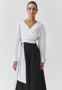 Witte wikkelblouse met lange mouwen en een diepe V-hals, gecombineerd met een zwarte rok. Soepele stoftextuur met een gestrikte taille-detail.