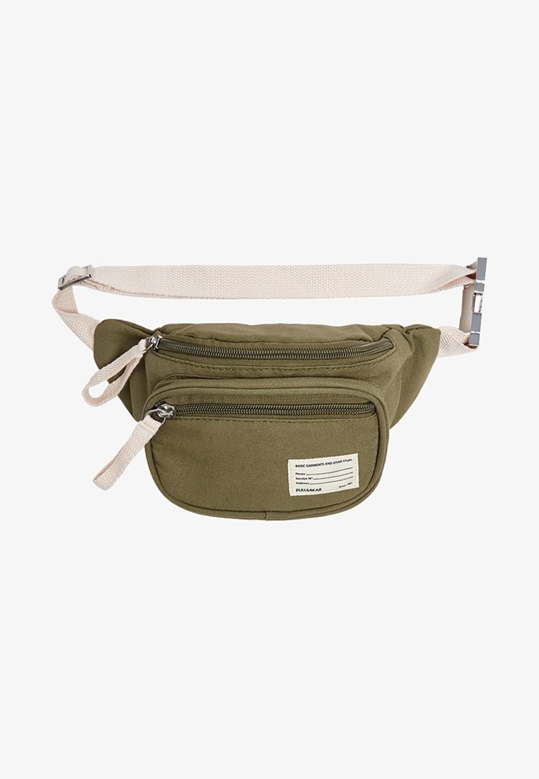 Sac banane en tissu vert olive avec deux compartiments zippés et une sangle beige réglable avec boucle métallique.