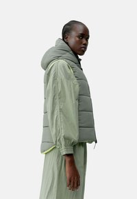 Lys grønn puffervest med hette, med et quiltet design og glidelås. Løs passform, akkompagnert av lysegule detaljer.