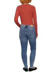 T-shirt long à manches ribbées en rust associé à un jean fitted bleu clair, avec des poches arrière et un détail en cuir sur la ceinture. Des ballerines noires.