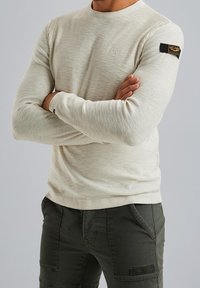 Chemise beige clair à manches longues avec un col rond et des détails côtelés. Comprend une étiquette noire sur la manche gauche et un tissu texturé.
