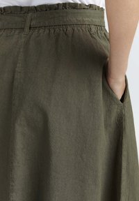 Olive green skirt with a textured finish, featuring a ruffled waistline and side pockets, shown from the back with a hand in a pocket.