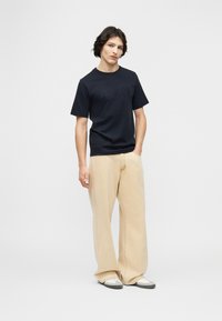 Νέος άνδρας στέκεται με το ένα χέρι στην τσέπη των μπεζ παντελονιών με φαρδιά μπατζάκια, φοράει μαύρο κοντομάνικο μπλουζάκι με λογότυπο και λευκά αθλητικά παπούτσια.