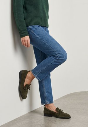 Person iført blå jeans, grøn sweater og olivengrønne ruskind loafers, der læner sig op ad en hvid væg med det ene ben bøjet.
