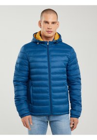 Egy férfi kék steppelt dzsekit visel sárga kapucninánnal és világoskék farmert, egyszerű világos háttér előtt áll.