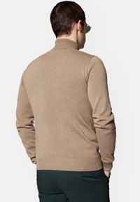 Beżowy sweter z dzianiny z wysokim kołnierzem, dopasowany krój, ściągacze przy mankietach i u dołu. Noszony z ciemnozielonymi spodniami. Wyświetlony widok z tyłu.