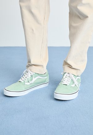Grønne semskede joggesko med hvite detaljer, rund tå, og hvite snørebånd. Kombinert med lyse bukser på et blått teppe.