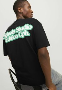 Camiseta de algodón negra con gran texto gráfico en blanco y verde menta en la parte posterior. Mangas cortas y ajuste relajado. El texto dice "Edición de Estudio Originals Cph."