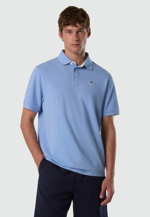 Hombre joven con pelo corto y rizado, vistiendo una camiseta polo azul claro con un pequeño logo redondo en el pecho y pantalones oscuros, de pie frente a un fondo liso.