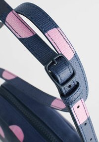 Jūras zila soma ar teksturētu apdari, kurai ir plata siksna ar rozā punktu rakstu; redzams regulējams sprādzes un rāvējslēdzēja detaļas.