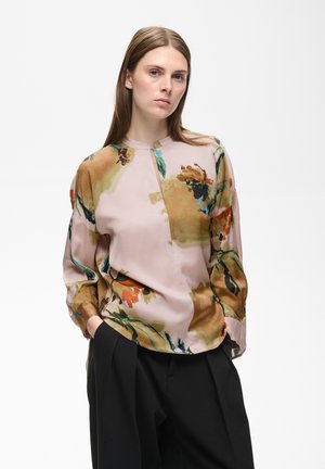 Kvinde med glat brun hår iført en lyserød bluse med lange ærmer med abstrakte blomsterprint og sorte bukser med vide ben, hænderne i lommerne.
