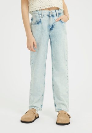 Jeans azzurri chiari a vita alta con una finitura sbiadita, vestibilità ampia, design a cinque tasche e un solo bottone in alto. Indossati con scarpe beige tipo slip-on.