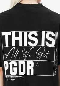 T-shirt in cotone nero con una grande grafica testuale bianca sul retro. Il testo include "QUESTO È", "Tutto ciò che abbiamo" e "PGDR."