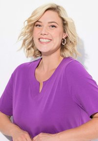 Fialový top s krátkým rukávem a výstřihem do V, hladká textura a volný střih. Modelka se usmívá s blond vlasy po ramena.