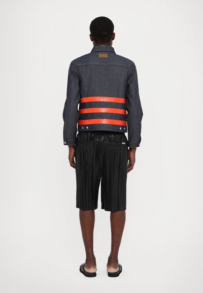 Veste en denim avec un design à rayures rouge et noire au niveau de la taille, associée à un short noir plissé et des sandales. Vue de dos.