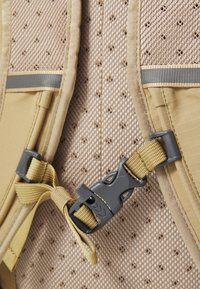 Beige fabric backpack section with mesh texture, gray accent straps, and a plastic clip. Features a patterned backing with small holes.