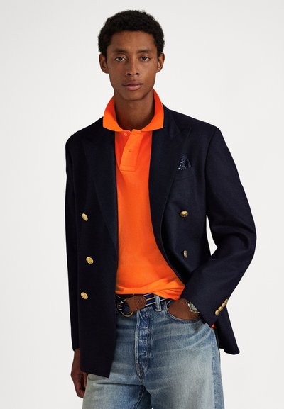 Blazer croisé bleu marine avec boutons dorés sur un polo orange vif. Porté avec un jean bleu clair et une ceinture marron, agrémenté d'un mouchoir de poche.