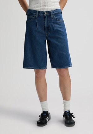 Άτομο που φοράει μακριά μπλε τζιν σορτς, λευκές κάλτσες, μαύρα και λευκά αθλητικά παπούτσια και μια λευκή αμάνικη μπλούζα φορεμένη μέσα, στέκεται μπροστά σε απλό φόντο.