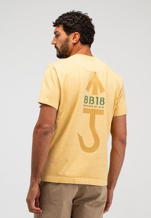 Man met krullend haar en baard, draagt een geel T-shirt met de opdruk "BB18 Butcher of Blue" en een grote gestileerde pijl en haak graphic op de achterkant.