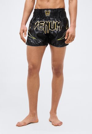 Άνδρας που φοράει μαύρα και χρυσά αθλητικά σορτς με τη σήμανση Venum και σχέδιο με φολίδες φιδιού, όρθιος ξυπόλητος μπροστά σε απλό φόντο.