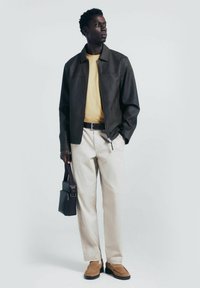 Giacca nera in finta pelle, t-shirt gialla, pantaloni crema, mocassini in suede marroni, mentre tiene una borsa scura. Design semplice, silhouette aderente.